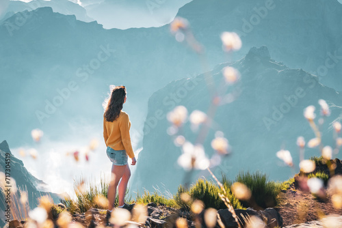 Female tourist stands on the windy edge of a deep, cloud-covered valley and enjoys the breathtaking panoramic view of the mountain landscape. Pico do Arieiro, Madeira Island, Portugal, Europe.