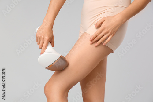 Young woman using photoepilator on legs against grey background