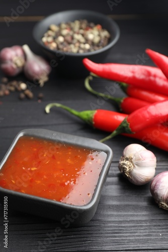 Spicy chili sauce, garlic, peppers and peppercorns on black wooden table, closeup