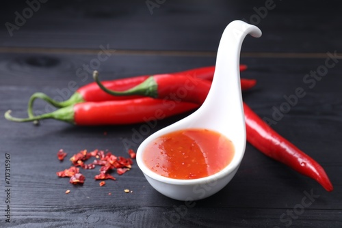 Spicy chili sauce in spoon and peppers on black wooden table, closeup