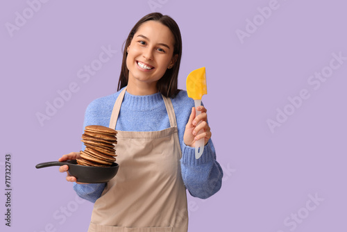 Beautiful young woman holding frying pan with tasty pancakes on lilac background