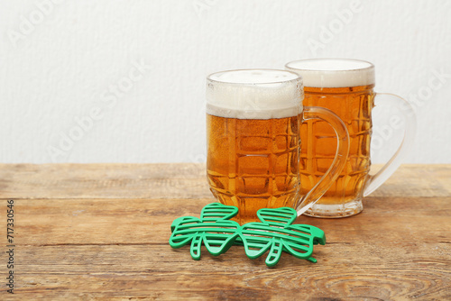 Mugs of beer and plastic eyeglasses on wooden table against white background. St. Patrick's Day celebration