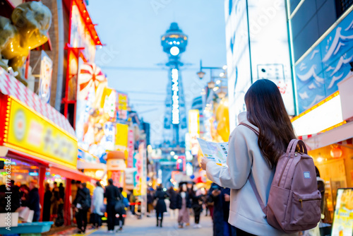 Asian female tourist Traveling and having fun. And she was taking photo with a cell phone camera at Night street with many restaurant around Tsutenkaku Tower in Shinsekai district of Osaka, Japan.