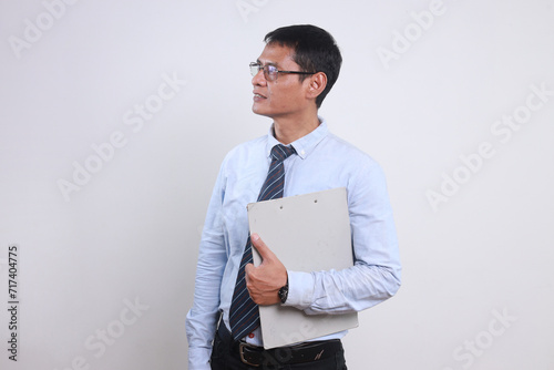 Adult Asian businessman wear glasses and formal office clothes, holding clipboard while looking away 