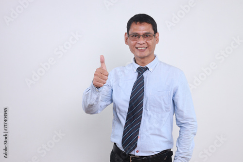 Happy young Asian businessman wear glasses and light blue shirt with tie, showing thumb up gesture over white background