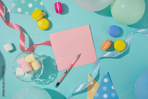 A pencil sits next to a pink card. Marshmallows, ribbons, balloons and macarons are decorated around the blue background. Free space to design birthday wishes.