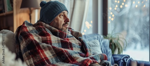 Man shivers at home in winter due to broken thermostat, wrapped in woolen plaid, sitting on sofa indoors. photo