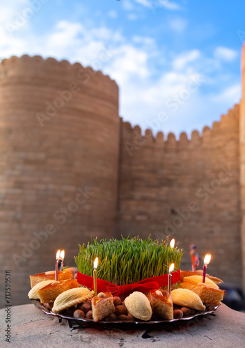Novruz celebration festive tray or plate with green wheat grass and traditional national pastry - pakhlava, shekerbura and candles with Baku old city in the background 