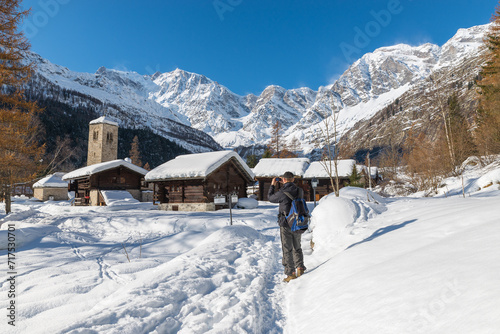 Hiker on vacation photographs a scenic winter landscape with snow in european Alps. Wonderful traditional mountain village of Macugnaga (Staffa - Pecetto), Italy, important ski resort with Monte Rosa