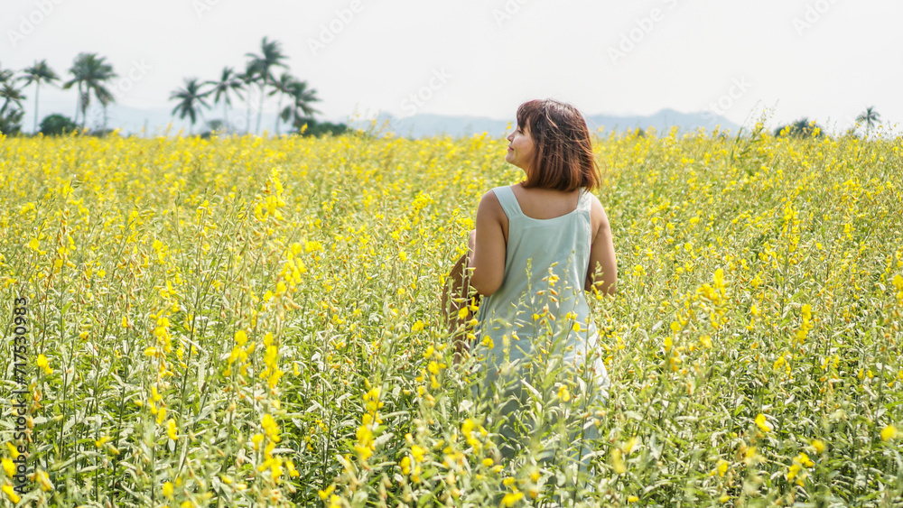Asian woman standing in beautiful yellow sunn hemp flower field with smiling happy face while travel outdoor. Female have freedom time garden with blue sky background. Concept of nature moment. Asian woman standing in beautiful yellow sunn hemp flower field with smiling happy face while travel outdoor. Female have freedom time garden with blue sky background. Concept of nature moment.