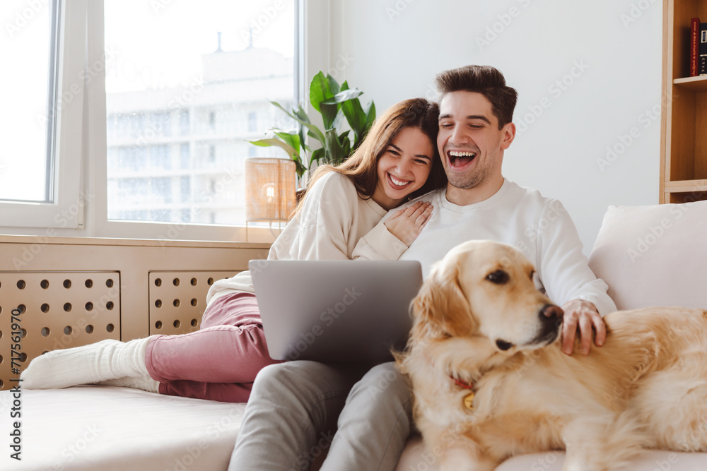 Smiling handsome boyfriend and girlfriend sitting on sofa, hugging watching video laughing Smiling handsome boyfriend and girlfriend sitting on sofa, hugging watching video laughing