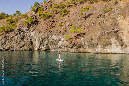 Aerial shot of single woman on paddle board move through calm sea water along rocks. Stand up paddle board in amazing scenery