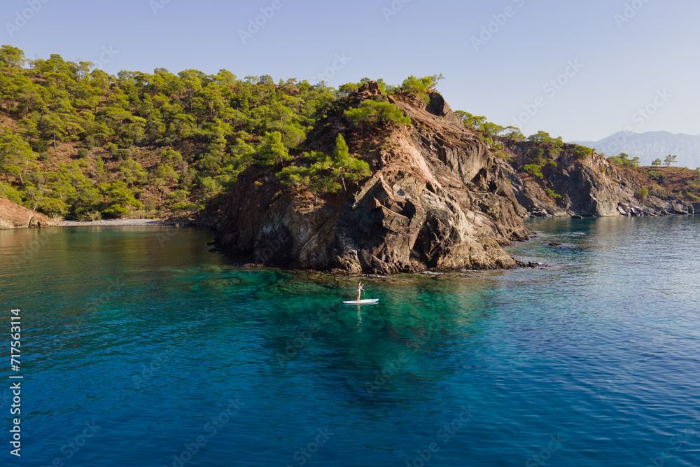 Aerial view of woman paddling a stand-up paddleboard or SUP board on a calm sea along rocky shore Aerial view of woman paddling a stand-up paddleboard or SUP board on a calm sea along rocky shore