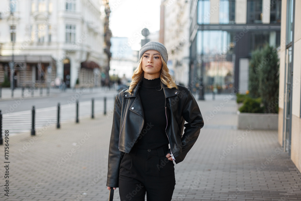 Fototapeta premium Fashionable beautiful young woman in stylish casual clothes with a knitted hat and fashion rock black leather jacket with a sweater walks in the city