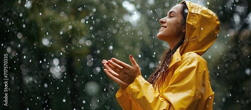 Horizontal side view image of a joyful young woman smiling wearing yellow raincoat during the rain in the nature Pretty female looking up and catching the rain drop with hands outdoors in the p