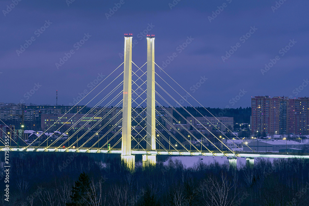 Fototapeta premium Night view of the silhouette of the illuminated Arkhangelsk bridge in Cherepovets against the background of lights of residential high-rise buildings.