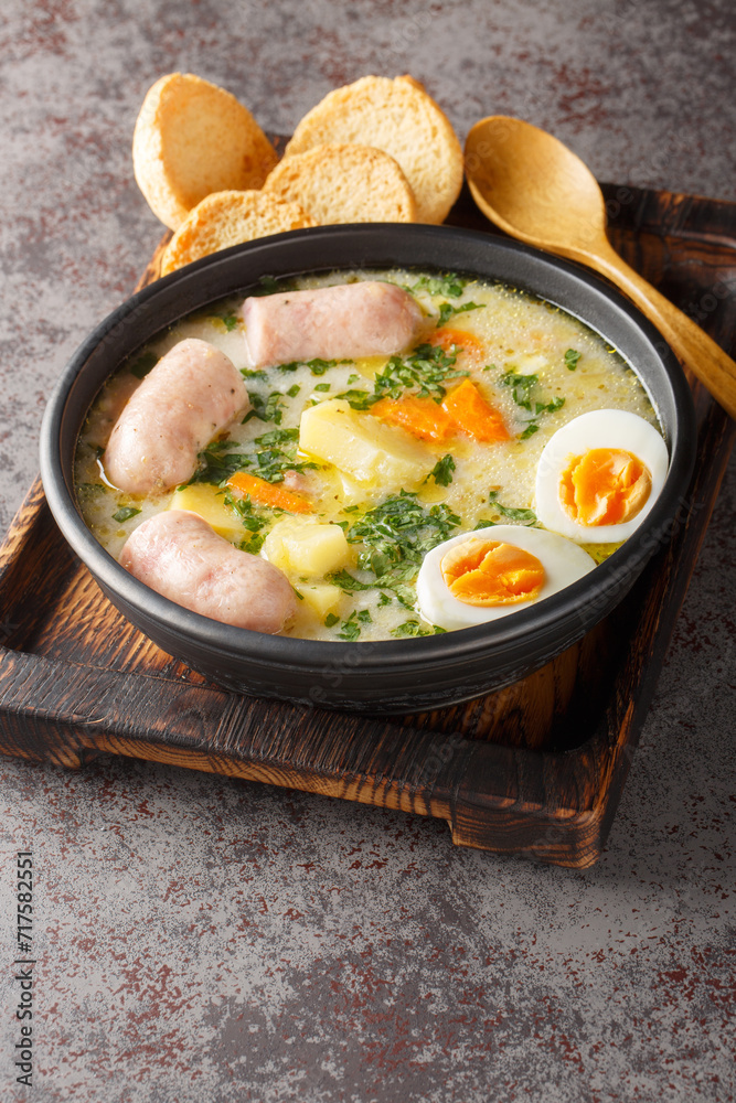 Authentic zurek a hearty Polish sour rye soup with rich white sausage and fresh veggies close-up in a bowl on the table. Vertical