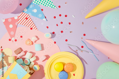 Marshmallow, macaron, colored candles, triangular streamers, birthday hats, balloons and confetti on a pastel purple background. Colorful birthday party.