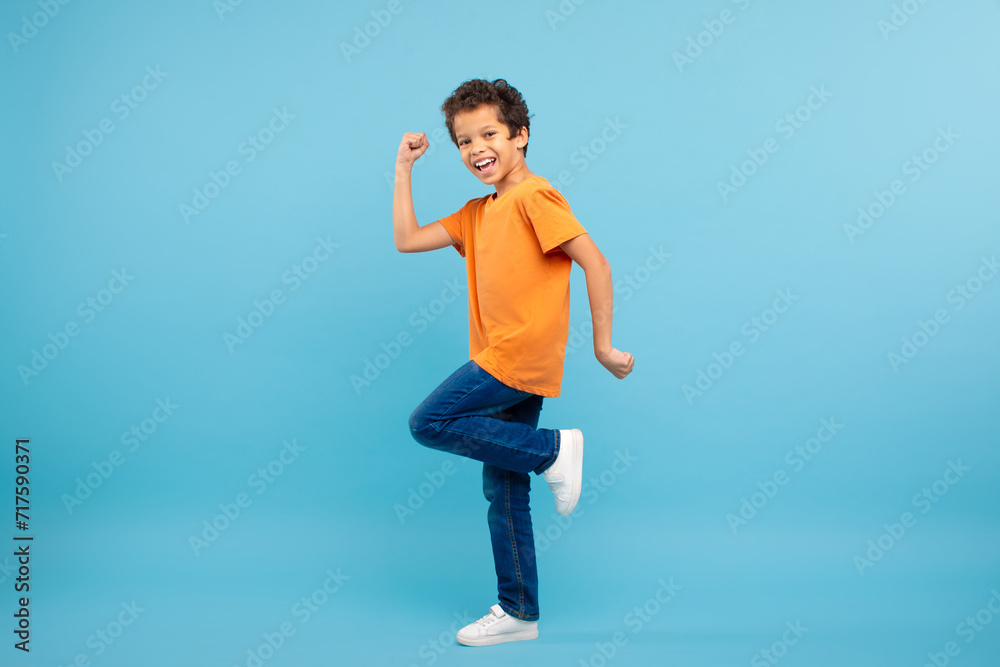 Joyful schoolboy in orange t-shirt clenching fist, blue background Joyful schoolboy in orange t-shirt clenching fist, blue background