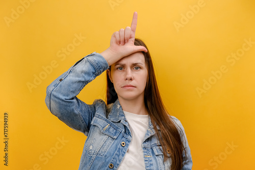Portrait of upset disappointed young caucasian woman showing loser gesture with hand on forehead, wearing denim jacket, posing isolated over yellow color background wall in studio. Failure concept photo