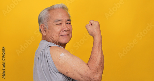 Asian old man showing adhesive plaster bandage on shoulder after vaccination. Isolated on yellow background in the studio photo