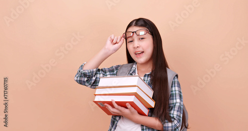 Cute school girl is carrying a school bag and holding a textbook. Isolated on brown background in studio.