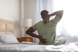 african man sitting on bed touching sore back and neck