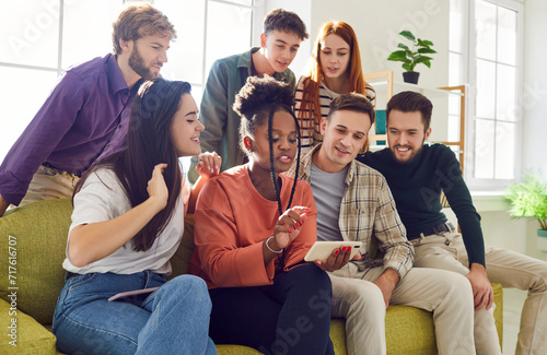 Group of friends use mobile phone together to view photos, videos or search Internet. Multiracial young people sitting on sofa at home and looking at smartphone screen. Best pals gathering concept.