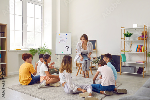 Group of elementary school students sitting on floor listening to teacher in classroom. School children sitting cross legged in circle around smiling female teacher listening attentively photo