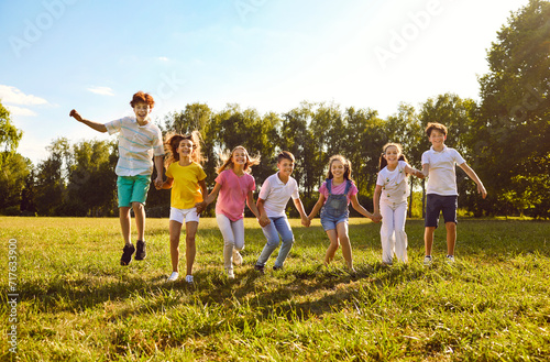 Group of a smiling kids friends jumping with hands up on green grass in the park standing in a line. Happy children having fun together outdoors on a sunny summer day in casual clothes in the camp.