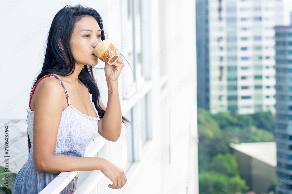 A woman in a summer dress drinks from a teacup on the balcony of an apartment building in a residential area A woman in a summer dress drinks from a teacup on the balcony of an apartment building in a residential area
