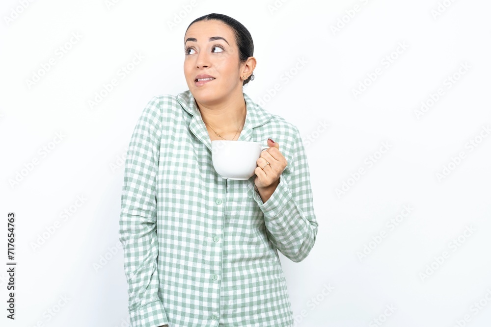 Amazed Beautiful young woman wearing green plaid pyjama and holding a cup bitting lip and looking tricky to empty space. Amazed Beautiful young woman wearing green plaid pyjama and holding a cup bitting lip and looking tricky to empty space.