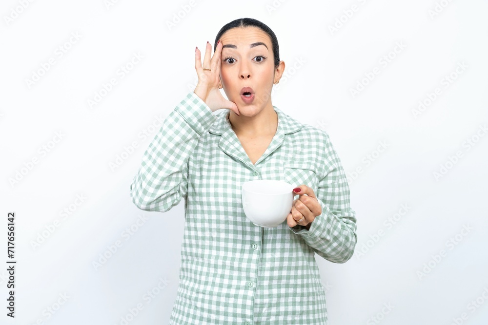 Beautiful young woman wearing green plaid pyjama and holding a cup with scared expression, keeps hands on head, jaw dropped, has terrific expression. Omg concept Beautiful young woman wearing green plaid pyjama and holding a cup with scared expression, keeps hands on head, jaw dropped, has terrific expression. Omg concept