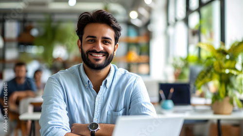 A cheerful and prosperous businessman is depicted in this portrait, grinning and gazing at the camera. He seems pleased with his accomplishments. The man is working on a laptop inside an office buildi photo