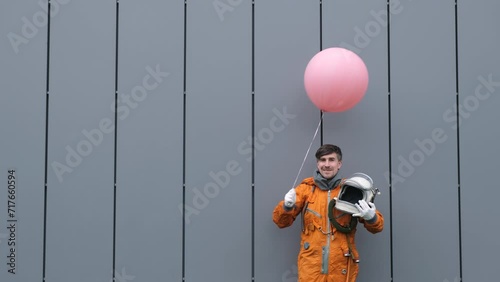 Happy astronaut wearing space suit takes space helmet off and smile while holding pink helium balloon outdoors. Surreal concept photo
