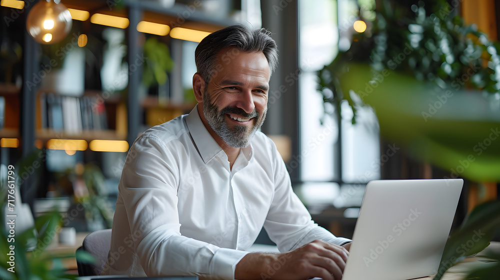 A smiling adult businessman who is using a laptop while seated at his desk. A satisfied and active middle-aged businessman, CEO, and manager who works in an office using corporate technology A smiling adult businessman who is using a laptop while seated at his desk. A satisfied and active middle-aged businessman, CEO, and manager who works in an office using corporate technology