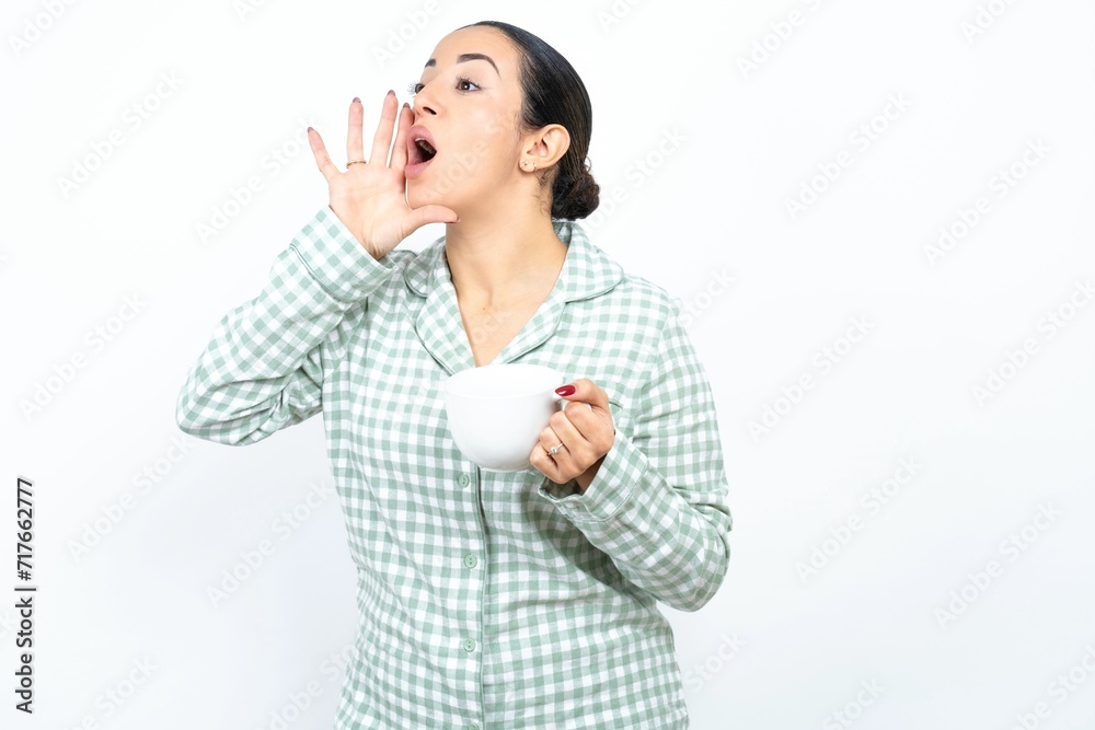 Beautiful young woman wearing green plaid pyjama shouting and screaming loud to side with hand on mouth. Communication concept. Beautiful young woman wearing green plaid pyjama shouting and screaming loud to side with hand on mouth. Communication concept.