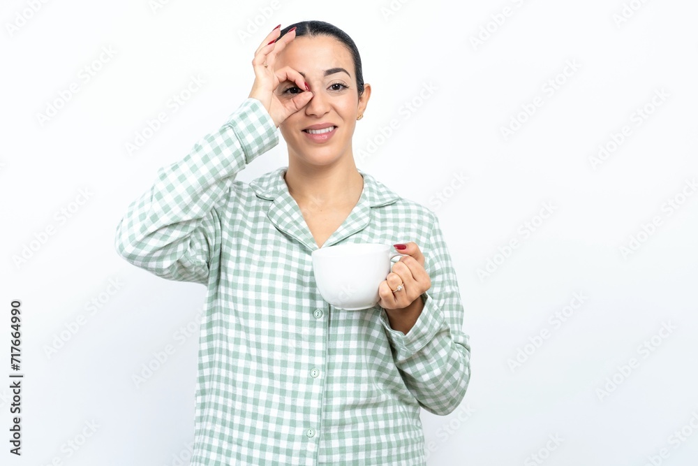Beautiful young woman wearing green plaid pyjama doing ok gesture with hand smiling, eye looking through fingers with happy face. Beautiful young woman wearing green plaid pyjama doing ok gesture with hand smiling, eye looking through fingers with happy face.