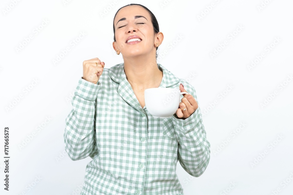 Beautiful young woman wearing green plaid pyjama being excited for success with raised arms and closed eyes celebrating victory. Winner concept. Beautiful young woman wearing green plaid pyjama being excited for success with raised arms and closed eyes celebrating victory. Winner concept.