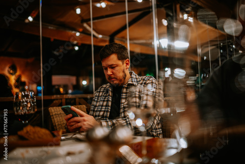 Malaga, Spain - January, 21, 2024 -  man in a plaid shirt looking intently at his smartphone in a restaurant setting with blurred lights... © Raivo