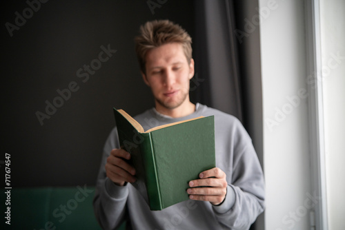 A focused young man wearing a casual grey sweater is deeply absorbed in reading a green hardcover book while seated comfortably by a window sill.