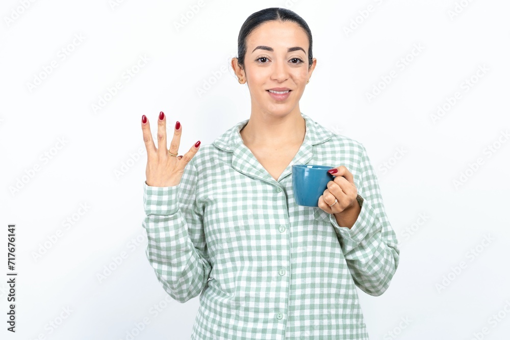 Beautiful young woman wearing green plaid pyjama and holding a cup smiling and looking friendly, showing number four or fourth with hand forward, counting down Beautiful young woman wearing green plaid pyjama and holding a cup smiling and looking friendly, showing number four or fourth with hand forward, counting down