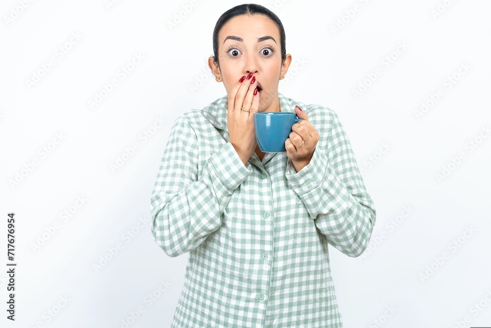 Beautiful young woman wearing green plaid pyjama and holding a cup keeps hands on mouth, looks with eyes full of disbelief, being puzzled with amount of work Beautiful young woman wearing green plaid pyjama and holding a cup keeps hands on mouth, looks with eyes full of disbelief, being puzzled with amount of work