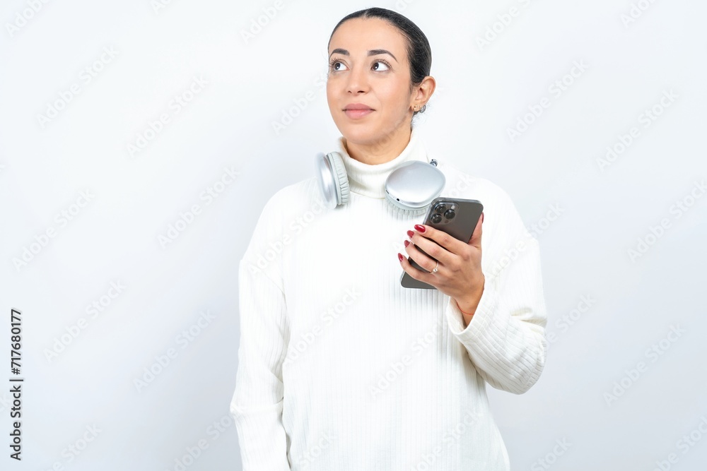 Young beautiful woman wearing white turtleneck sweater holds mobile phone uses high speed internet and social networks has online communication. Modern technologies concept Young beautiful woman wearing white turtleneck sweater holds mobile phone uses high speed internet and social networks has online communication. Modern technologies concept