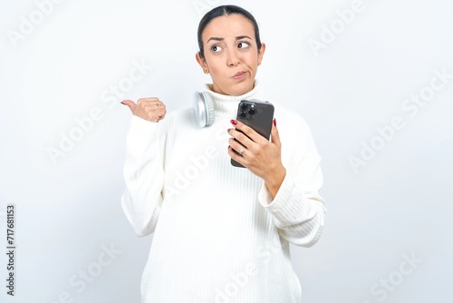 Young beautiful woman wearing white turtleneck sweater points thumb away and shows blank space aside, holds mobile phone for sending text messages.