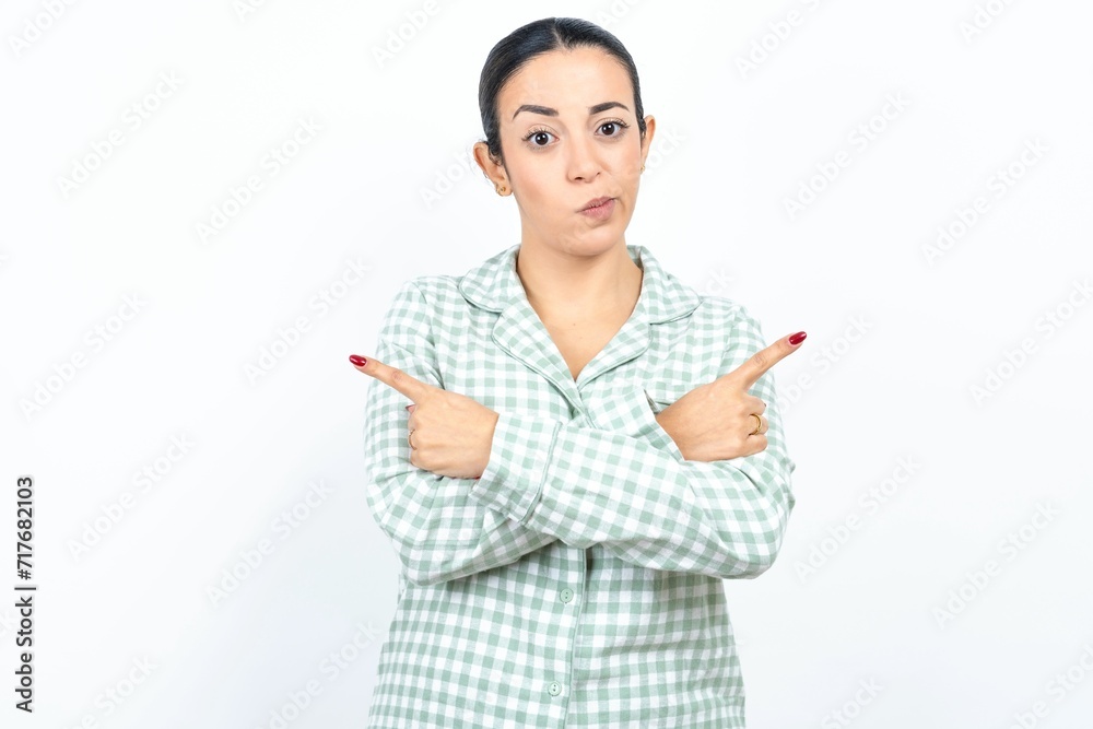 Serious Beautiful young woman wearing green plaid pyjama and holding a cup crosses hands and points at different sides hesitates between two items. Hard decision concept Serious Beautiful young woman wearing green plaid pyjama and holding a cup crosses hands and points at different sides hesitates between two items. Hard decision concept