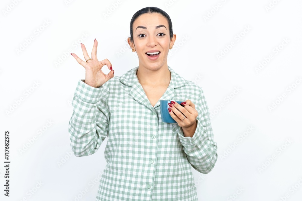 Beautiful young woman wearing green plaid pyjama and holding a cup hold hand arm okey symbol toothy approve advising novelty news Beautiful young woman wearing green plaid pyjama and holding a cup hold hand arm okey symbol toothy approve advising novelty news