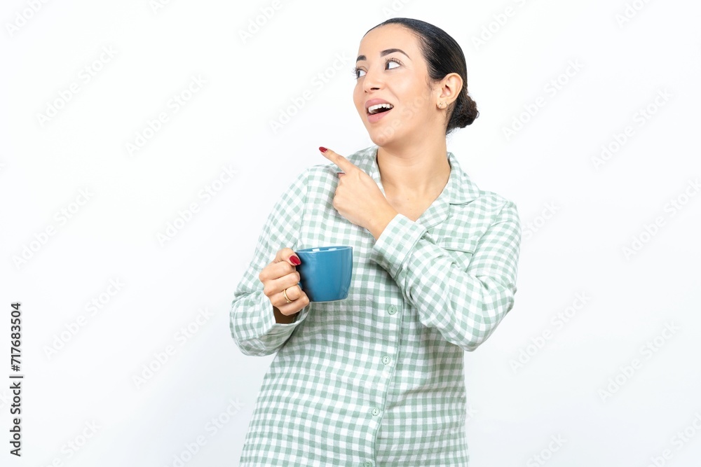 Beautiful young woman wearing green plaid pyjama and holding a cup glad cheery demonstrating copy space look novelty Beautiful young woman wearing green plaid pyjama and holding a cup glad cheery demonstrating copy space look novelty