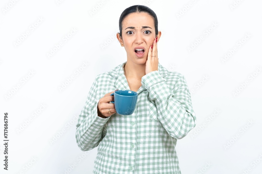 Upset Beautiful young woman wearing green plaid pyjama and holding a cup touching face with two hands Upset Beautiful young woman wearing green plaid pyjama and holding a cup touching face with two hands