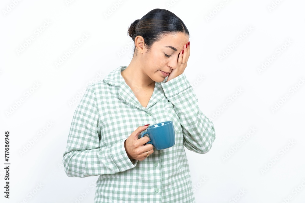 Beautiful young woman wearing green plaid pyjama and holding a cup with sad expression covering face with hands while crying. Depression concept. Beautiful young woman wearing green plaid pyjama and holding a cup with sad expression covering face with hands while crying. Depression concept.